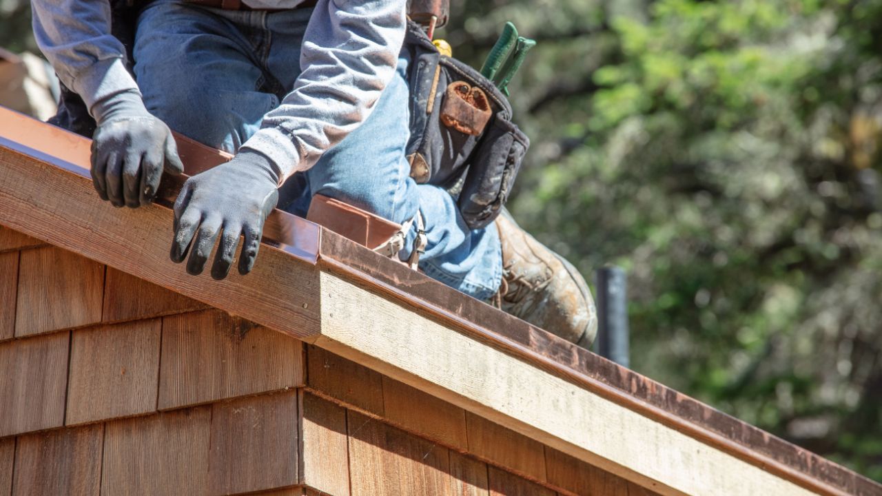 A person in gloves and jeans works on a wooden roof corner, surrounded by greenery, holding tools for construction. Installing the ice and water shield.