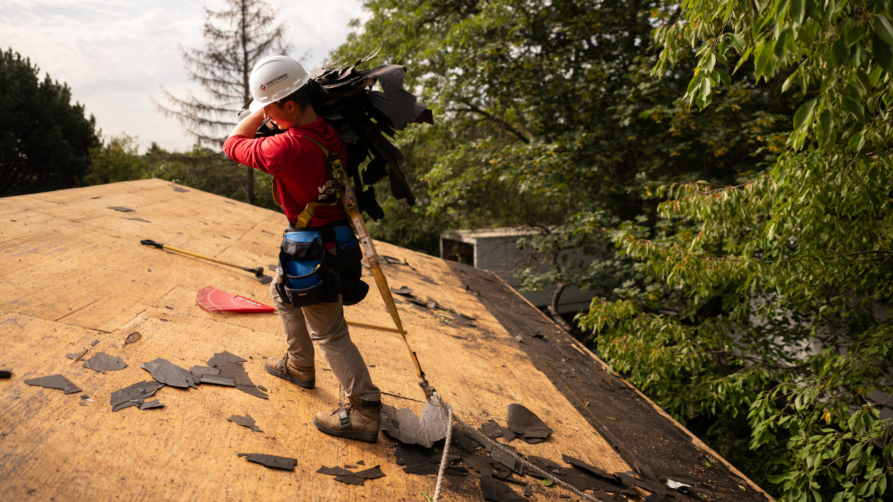 Person on roof, wearing a safety harness and helmet, carries shingles. Surroundings include scattered debris, trees, and safety equipment such as a shovel.