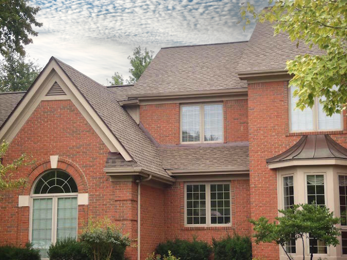 A two-story red brick house with gabled roof, large windows, and green bushes, surrounded by trees and under a partially cloudy sky.