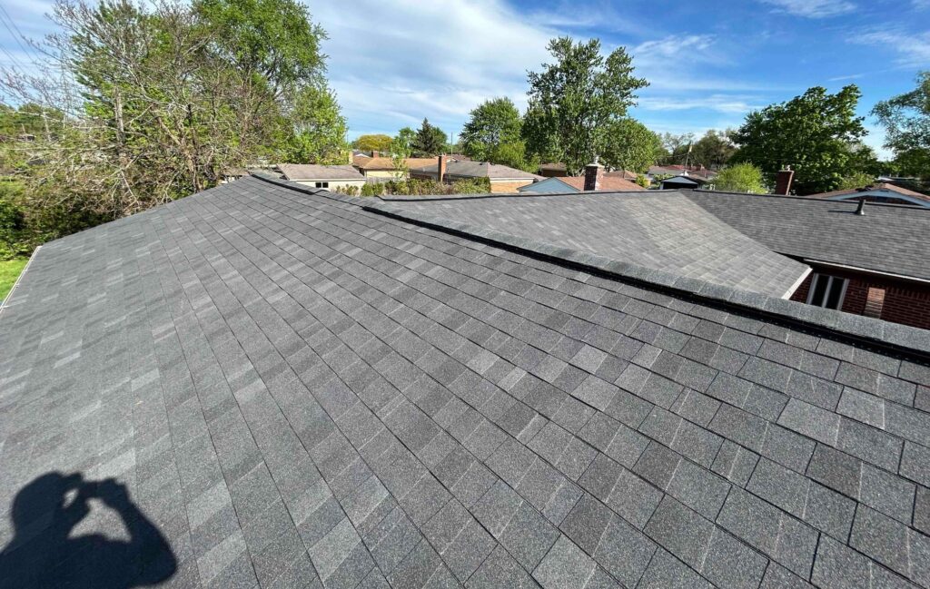 Gray shingled rooftops stretch across suburban houses with lush trees and blue sky. A person's shadow is visible on the roof.