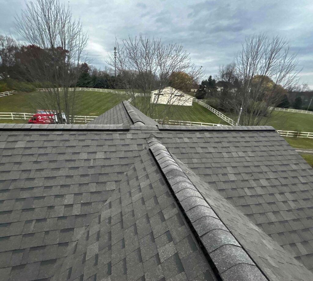 Shingle roof foreground with sprawling grassy field, white fences, and trees in background. Red vehicle parked near a white building. Cloudy sky.