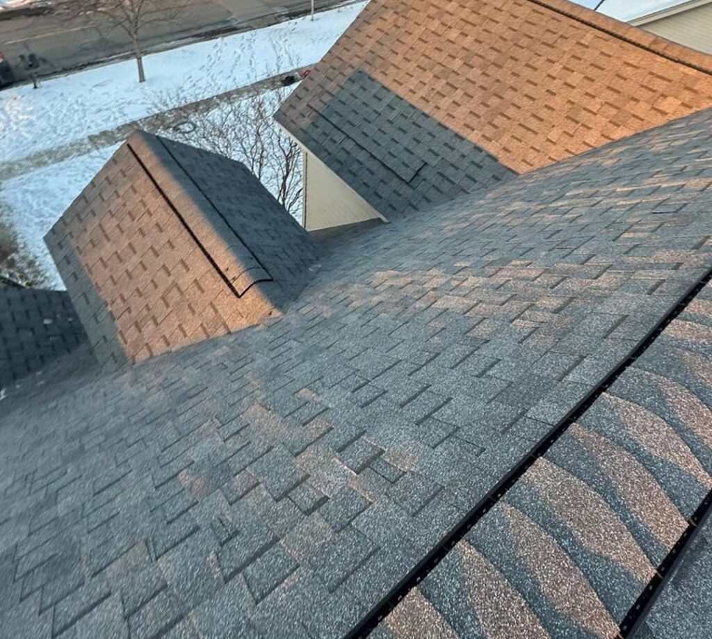 Asphalt-shingled rooftops in a suburban neighborhood, covered in snow. Bare trees and a residential street are visible in the background.