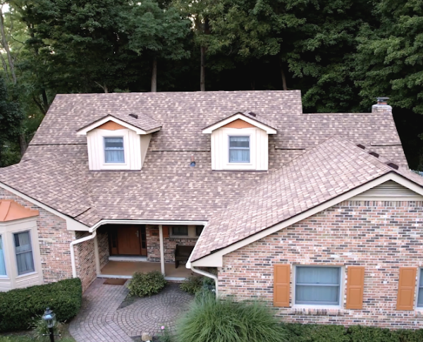 A brick house with a shingled roof, surrounded by trees. The entrance features decorative shrubs and a curved paved pathway.