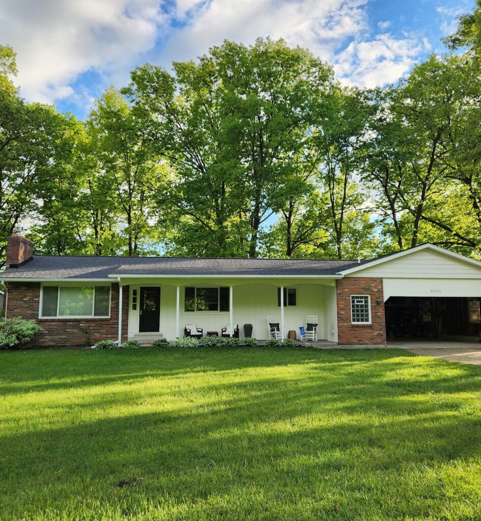 Single-story brick house with front lawn, surrounded by tall trees under a partly cloudy sky. Empty porch with chairs and pots.