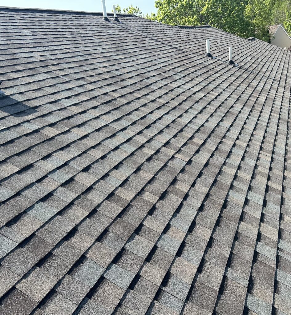 A textured, gray shingled roof with several white vent pipes beneath a clear blue sky, surrounded by green trees.